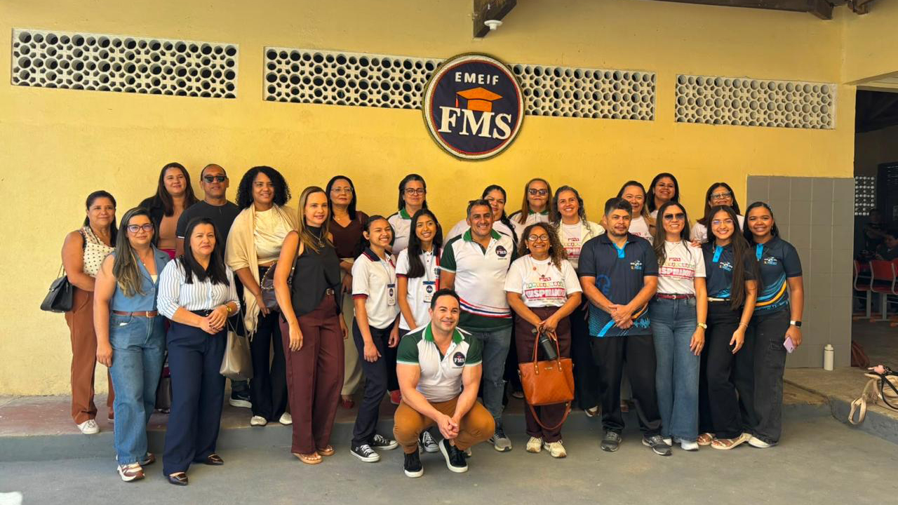 Um grupo multirracial de cerca de 25 pessoas, entre adultos e jovens, em trajes casuais, está de pé posando lado a lado. Acima deles, uma placa circular fixada em uma parede amarela exibe o texto “EMEIF FMS” e o desenho de um capelo laranja. O piso é cinza e a parede atrás do grupo conta com blocos vazados.