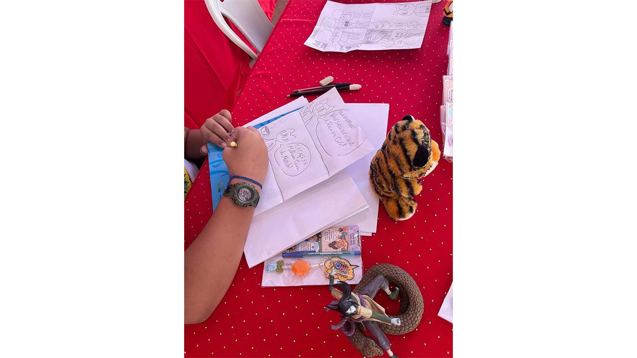 Uma criança usando um relógio preto está desenhando. O destaque é o papel com desenhos, no formato de história em quadrinhos, cercado por um tigre de pelúcia, canetas e um boneco articulado. O fundo resume-se à uma toalha vermelha com bolinhas brancas e a outros papéis.