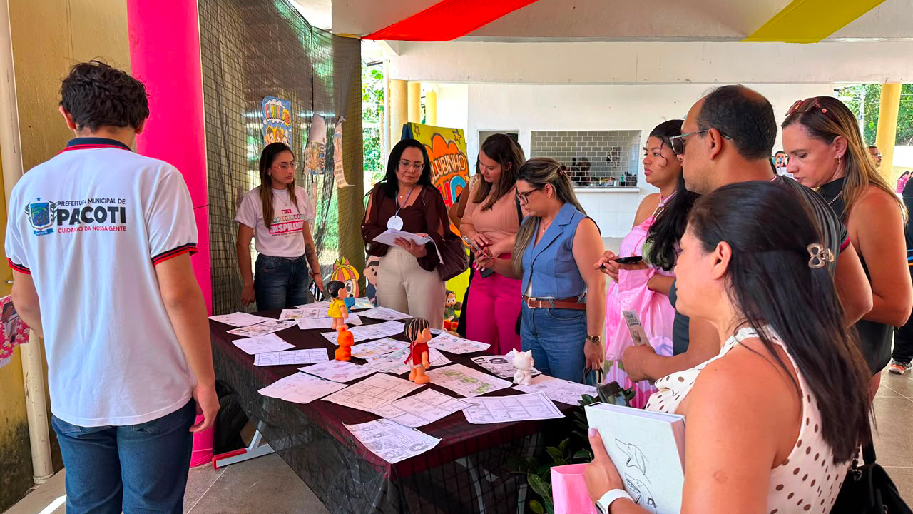 Um grupo multirracial de cerca de dez adultos, em pé, observa os itens dispostos sobre a mesa central, forrada com uma toalha escura. Sobre ela, há dezenas de páginas desenhadas e pequenos bonecos. O cenário inclui tecidos coloridos pendurados no teto e pilares brancos. De costas, um homem veste uma camiseta branca com os escritos "Pacoti".