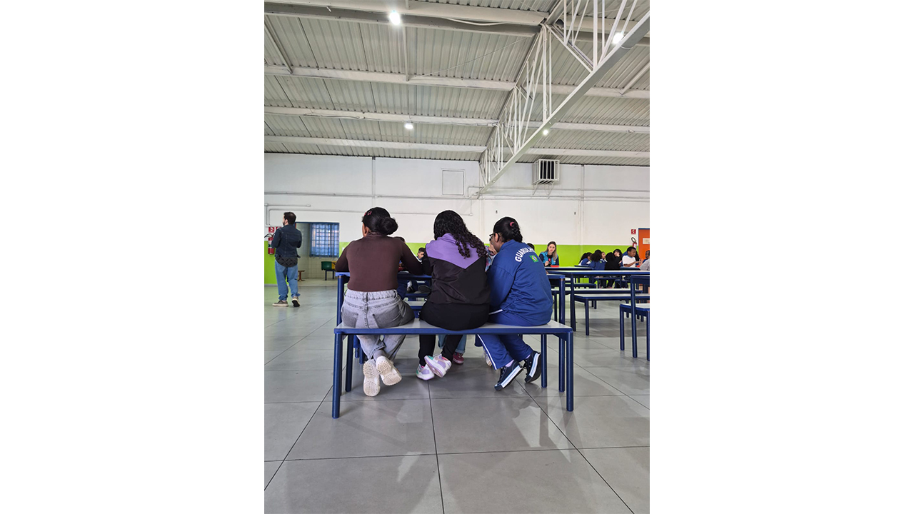Três jovens, sentadas de costas em um banco, conversam. O espaço é composto por conjuntos padronizados de mesas e bancos coletivos, nas cores branco e azul, além de paredes brancas com a metade inferior verde e teto com estrutura metálica.