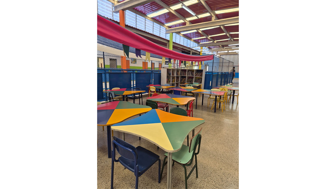 Espaço de convivência escolar com mesas com tampos pintados em triângulos coloridos e cadeiras também coloridas. Ao fundo, há estantes com livros, grades de proteção azuis, tela de proteção e teto metálico decorado com faixas de tecido rosa.