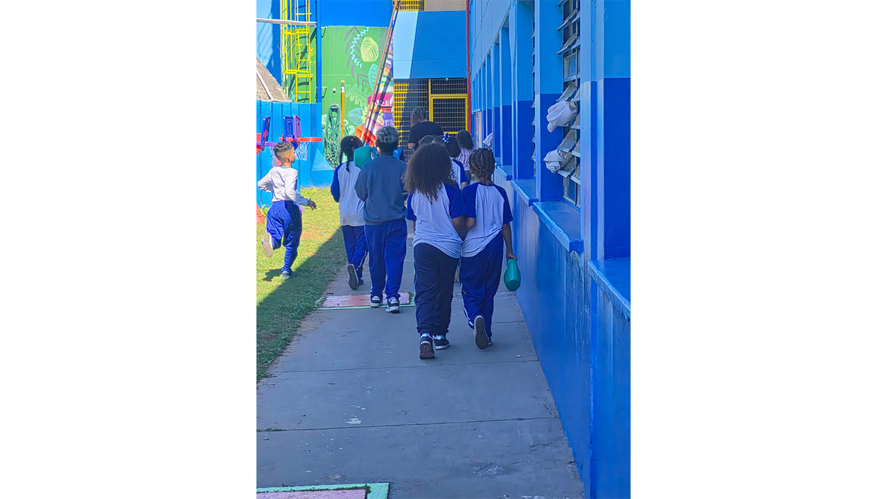 Um pequeno grupo multirracial de crianças caminha de costas, vestindo roupas semelhantes nas cores azul e branca; algumas seguram regadores de plástico verdes. Elas caminham por um estreito caminho de cimento, ao lado de uma longa construção azul, com janelas basculantes à direita, além de um gramado e um muro pintado à esquerda.
