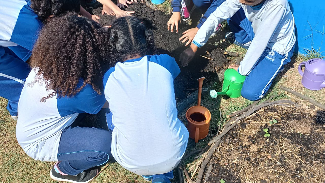 Crianças vestindo roupas semelhantes, nas cores azul e branca, estão agachadas e manuseiam a terra escura. Ao redor do grupo, estão regadores de plástico nas cores verde, lilás e marrom. As crianças estão na grama, em um canteiro delimitado por galhos secos e a base de uma parede azul à direita.