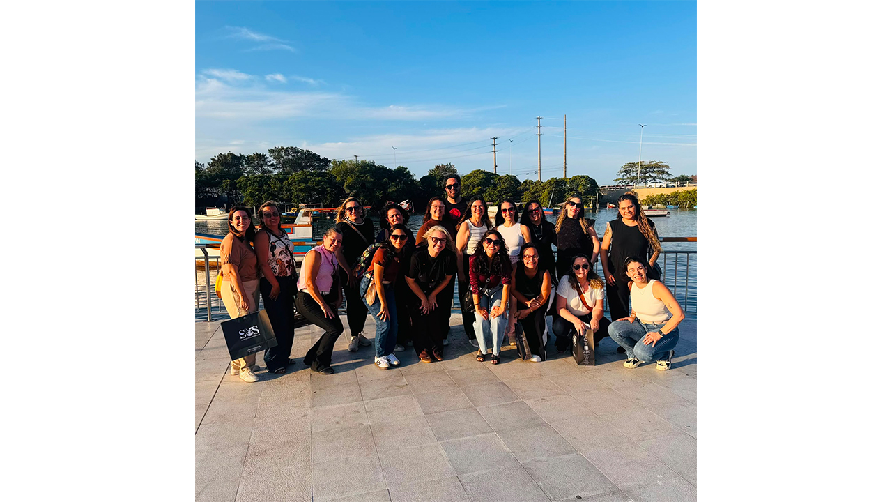 Um grupo multirracial de cerca de 19 pessoas posa para foto em uma área externa à beira da água. Algumas pessoas estão em pé e outras agachadas, usando roupas casuais de diversas cores. Ao fundo, aparecem embarcações, árvores e postes de iluminação.