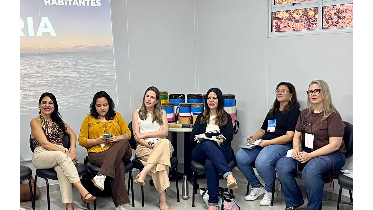 Seis mulheres estão sentadas em cadeiras pretas estofadas, organizadas em semi círculo, durante uma atividade. Algumas seguram celular, caderno ou caneta. Elas vestem roupas casuais e sociais. Ao fundo, há recipientes cilíndricos coloridos, pintados com faixas horizontais azuis, rosas e brancas, além de uma parede clara, parte de uma janela e uma tela de projeção ligada, onde é possível ver parte de uma fotografia de um corpo d'água.
