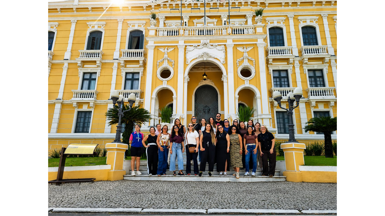 Área externa diante de um edifício histórico amarelo e branco, com um grupo de 21 adultos reunido na escadaria de acesso ao local. As pessoas posam lado a lado, algumas usando óculos escuros e roupas em tons variados. Ao fundo, destacam-se arcos, janelas, ornamentos, palmeiras e postes decorativos.