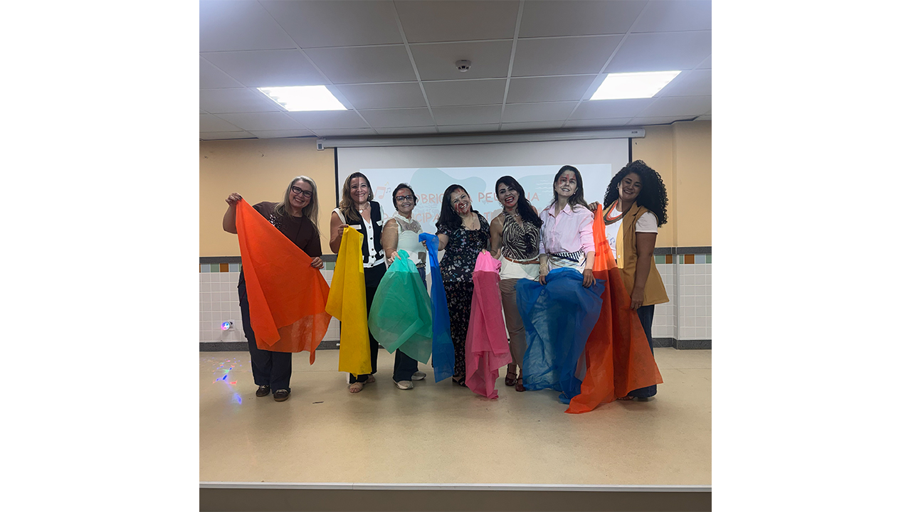 Grupo multirracial composto por sete mulheres posa lado a lado. Elas seguram tecidos coloridos em tons de laranja, amarelo, verde, azul e rosa. Elas estão em cima de um pequeno palco dentro de uma sala de paredes na cor salmão e piso claro. Ao fundo, há uma tela de projeção com texto parcialmente visível.