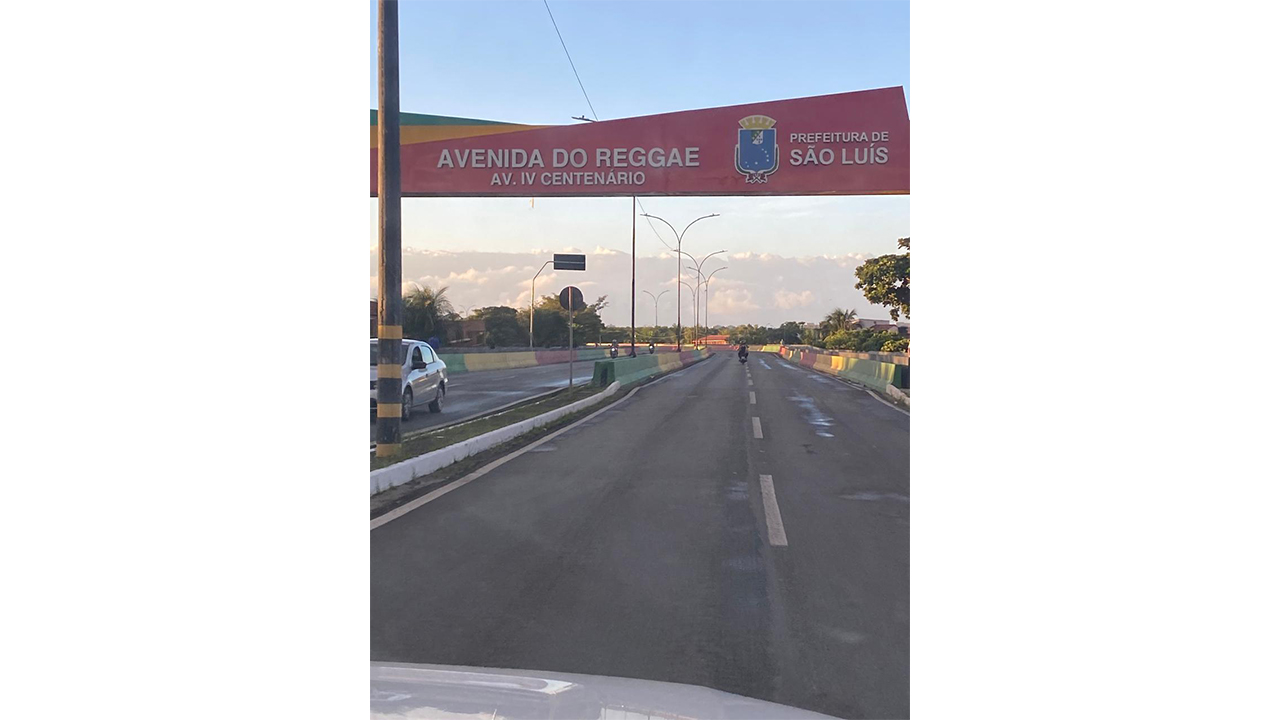 Em uma avenida, um portal vermelho exibe os textos “Avenida do Reggae”, “Av. IV Centenário” e “Prefeitura de São Luís”. Ao fundo, há muretas coloridas, postes e árvores.
