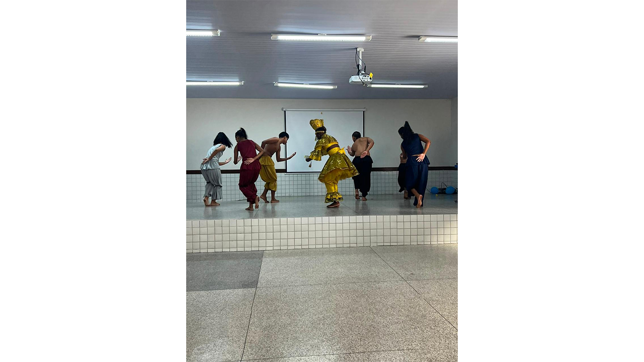 Grupo multirracial de seis pessoas realiza uma coreografia. O dançarino central veste trajes dourados e uma coroa, enquanto os demais usam roupas largas em tons escuros e terrosos. Eles dançam descalços sobre um palco de cerâmica branca, em uma sala fechada, com um telão de projeção desligado ao fundo.