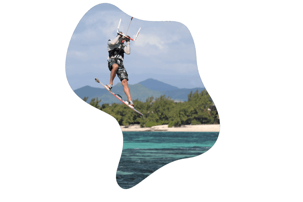 Um kitesurfista está no ar, pulando acima da água. A pessoa veste um short escuro na altura dos joelhos e uma camiseta branca de mangas compridas. A prancha conectada a seus pés tem a lateral vermelha e detalhes em branco. Uma corda amarrada à sua cintura a conecta à pipa, enquanto segura em uma estrutura de apoio branca com as duas mãos. A água do mar é azul-esverdeada, e há um homem na água, vestindo uma roupa preta. No plano de fundo, uma praia de areia branca ladeada por árvores verdes e montanhas baixas