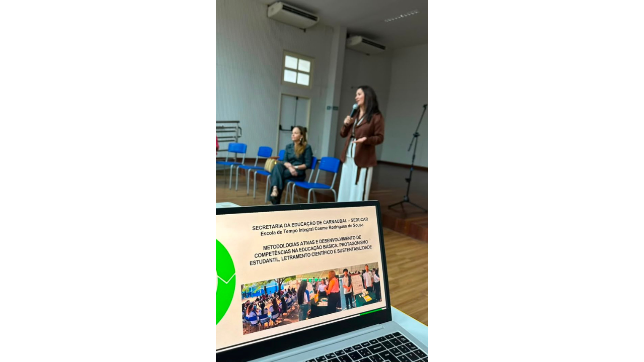 No centro de um auditório, uma mulher de blusa estampada preta e branca e calça clara fala ao microfone, enquanto pessoas sentadas na plateia a observam. À direita, um telão de projeção exibe fotografias coloridas, ao lado de uma mesa com um projetor e notebook. O espaço possui piso de madeira, cadeiras azuis e pretas enfileiradas, portas brancas e paredes claras com textura ripada.