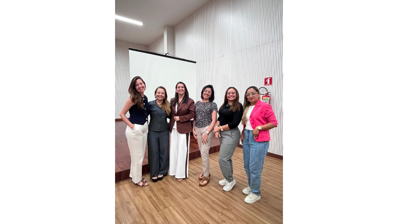 Um notebook exibe uma apresentação com fundo roxo e título "Secretaria Municipal de Educação de Guarulhos", enquanto, atrás dele e ao centro, uma mulher em pé, de blusa estampada, fala ao microfone. Outras duas mulheres, com roupas casuais, estão sentadas ao fundo em cadeiras azuais e a observam. O ambiente possui piso de madeira e parede clara com textura ripada.