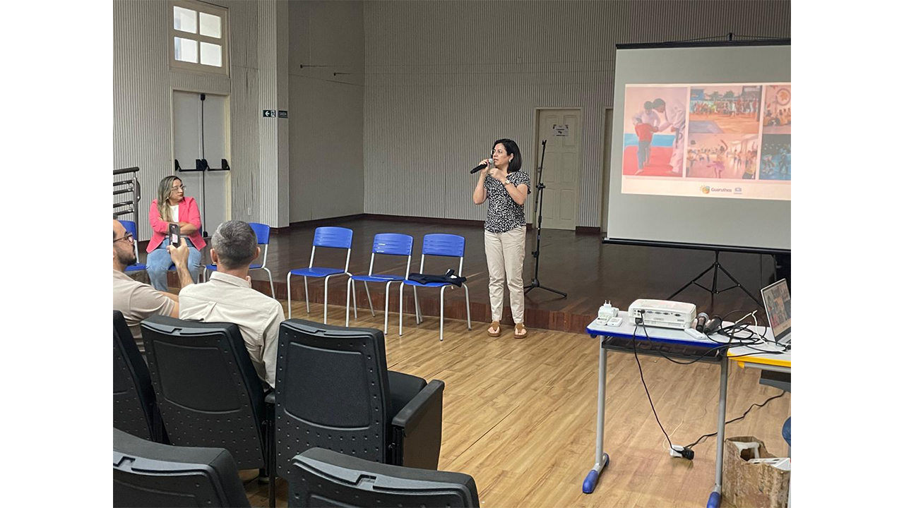 No centro de um auditório, uma mulher de blusa estampada preta e branca e calça clara fala ao microfone, enquanto pessoas sentadas na plateia a observam. À direita, um telão de projeção exibe fotografias coloridas, ao lado de uma mesa com um projetor e notebook. O espaço possui piso de madeira, cadeiras azuis e pretas enfileiradas, portas brancas e paredes claras com textura ripada.