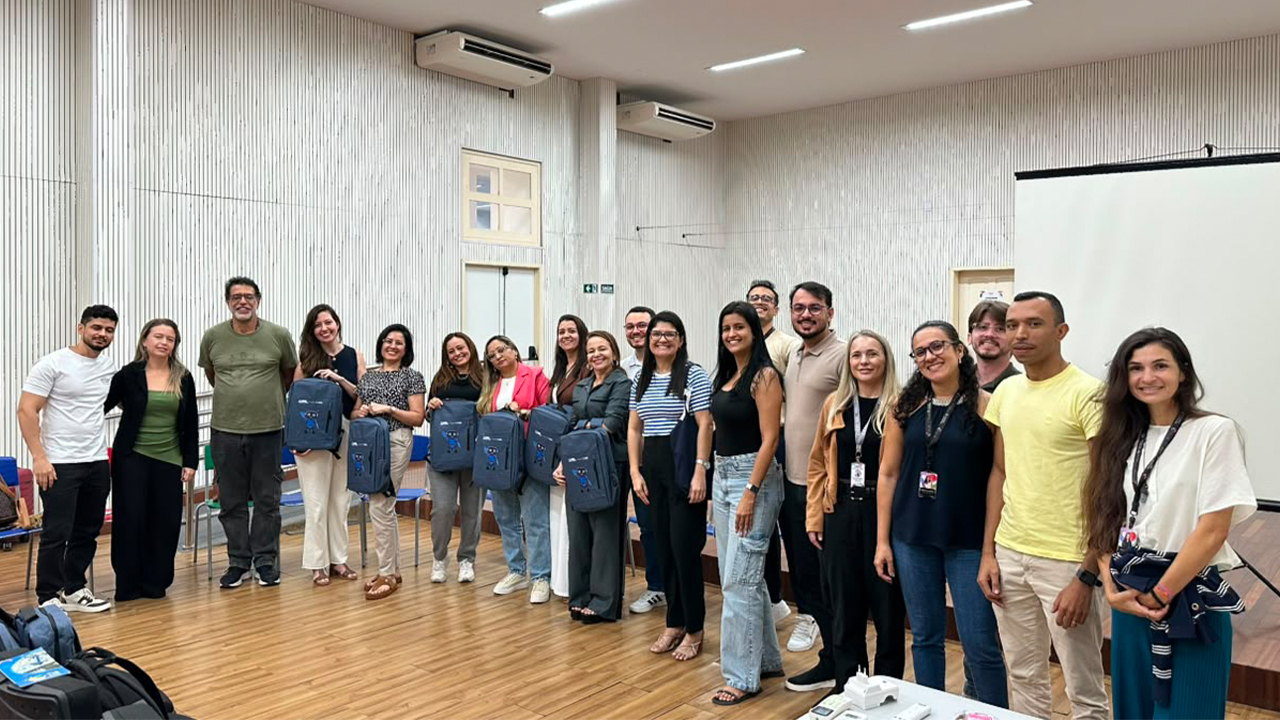 Grupo multirracial de cerca de vinte pessoas posa em pé para a foto, em um auditório iluminado, usando roupas casuais variadas. Algumas, no centro do grupo, seguram mochilas azuis idênticas à frente do corpo, com o desenho de um mascote impresso. O espaço conta com piso de madeira, um telão branco à direita e paredes brancas com frisos verticais, além de duas grandes máquinas de ar-condicionado próximas ao teto.