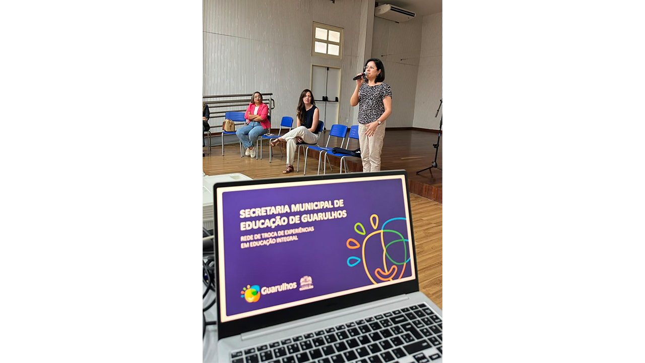 Um notebook exibe uma apresentação com fundo roxo e título "Secretaria Municipal de Educação de Guarulhos", enquanto, atrás dele e ao centro, uma mulher em pé, de blusa estampada, fala ao microfone. Outras duas mulheres, com roupas casuais, estão sentadas ao fundo em cadeiras azuais e a observam. O ambiente possui piso de madeira e parede clara com textura ripada.