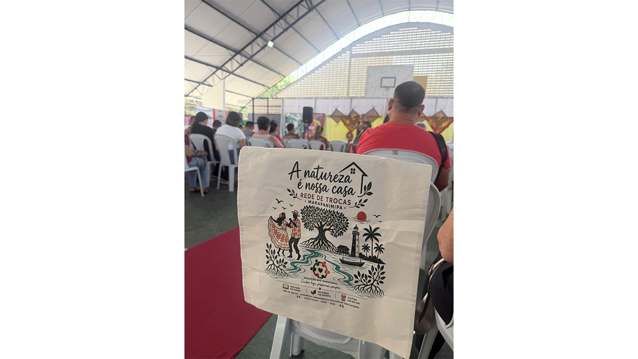 Uma ecobag pendurada na cadeira exibe uma ilustração de uma mulher e um homem dançando, uma casa, uma árvore, coqueiros e um farol, e o texto “A natureza é nossa casa — Rede de Trocas Marapanim/PA”. Ao fundo, diversas pessoas estão de costas, sentadas em cadeiras brancas, voltadas para um palco, próximo a uma tabela de basquete.
