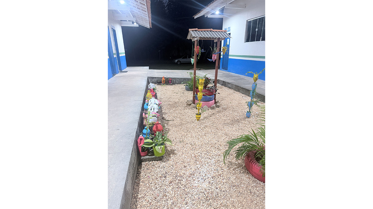 Jardim de pedras claras, com um poço decorativo feito de pneus coloridos - rosa, lilás e vermelho - empilhados e um pequeno telhado de zinco, adornado com vasos feitos de garrafas PET pintadas. Ao fundo, observam-se calçadas de cimento e a fachada branca e azul de um prédio, à noite.