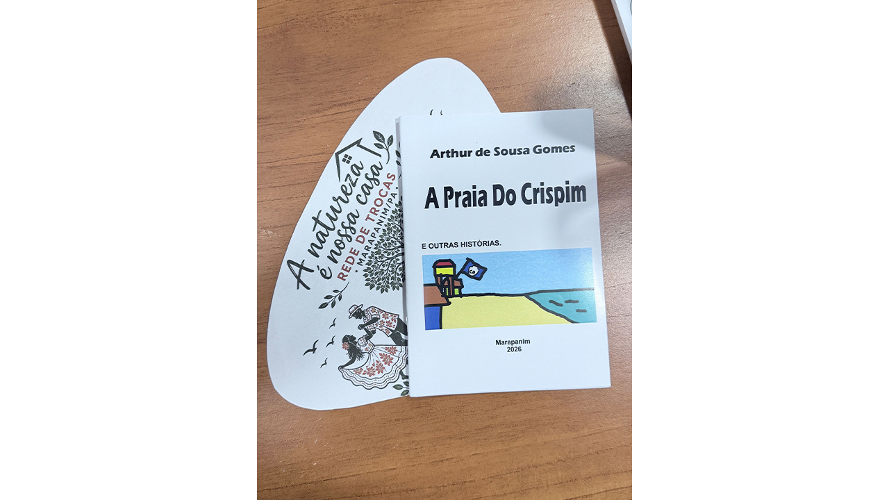 Um livreto intitulado “A Praia do Crispim e outras histórias”, de Arthur de Sousa Gomes, sobrepõe-se a um papel com o texto “A natureza é nossa casa — Rede de Trocas Marapanim/PA” e uma ilustração. O plano de fundo resume-se à textura lisa e marrom de uma mesa de madeira.