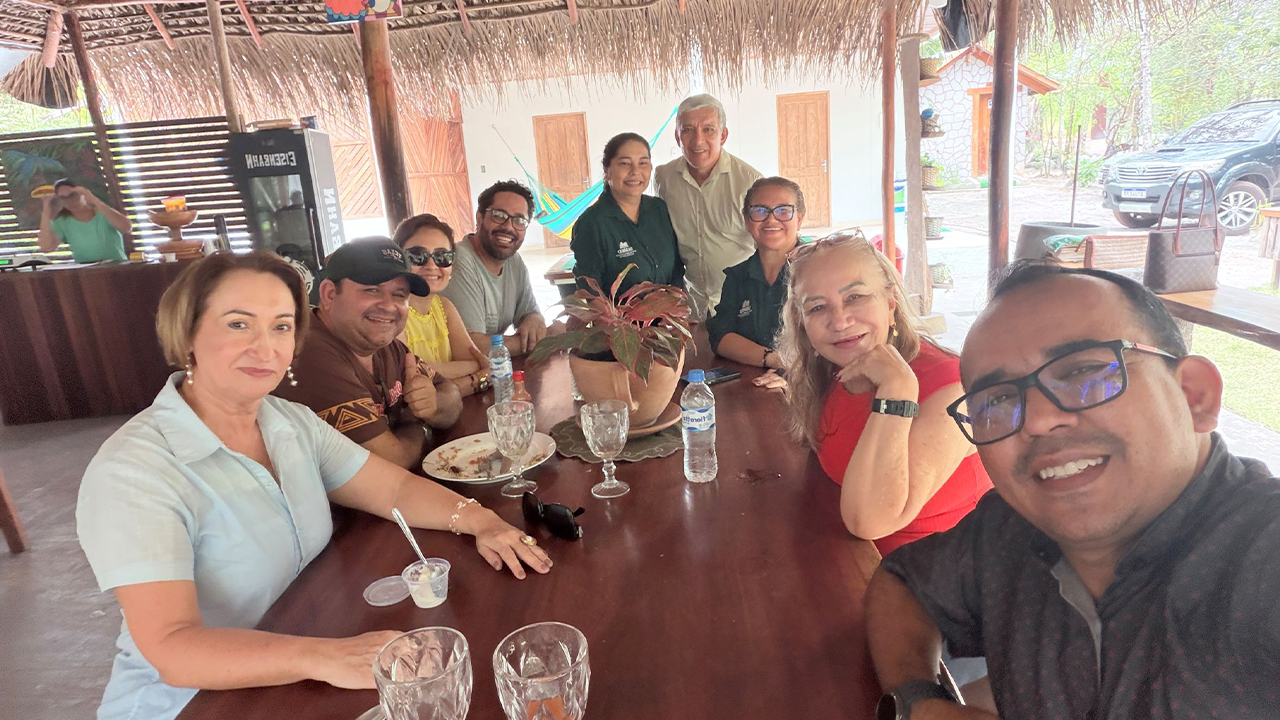 Grupo multirracial de nove pessoas, em trajes casuais, está sentado ao redor de uma mesa de madeira escura, onde repousam taças de vidro vazias, pratos e um pequeno vaso de planta. Ao fundo, vê-se a cobertura de palha do local, além de um carro azul estacionado à esquerda.