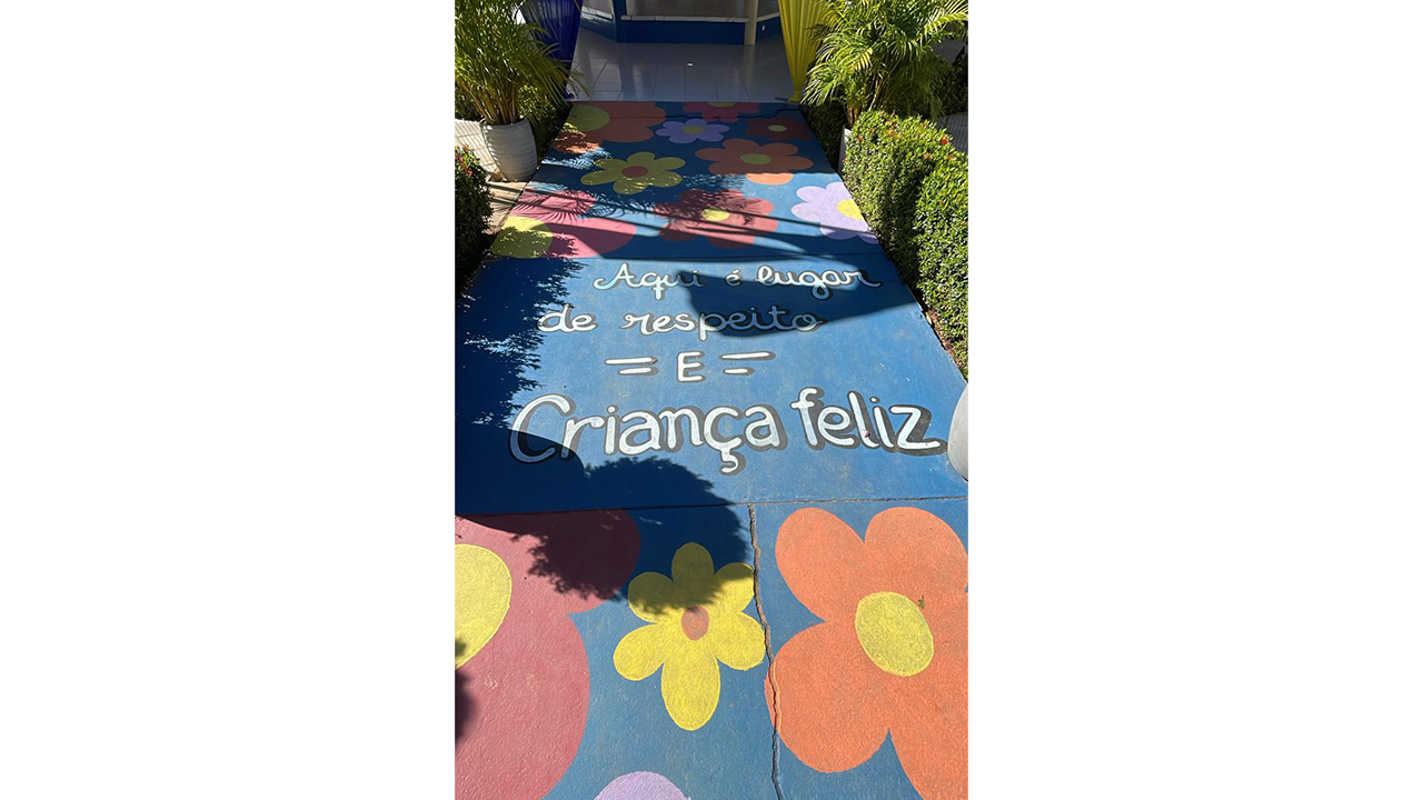 Um caminho no chão pintado de azul é decorado com desenhos de grandes flores em tons de vermelho, amarelo e roxo. No centro do trajeto, lê-se o texto escrito em branco: "Aqui é lugar de respeito e criança feliz". O cenário ao fundo mostra vasos com plantas verdes e cortinas de tecido em azul e amarelo que emolduram a entrada de uma estrutura coberta.