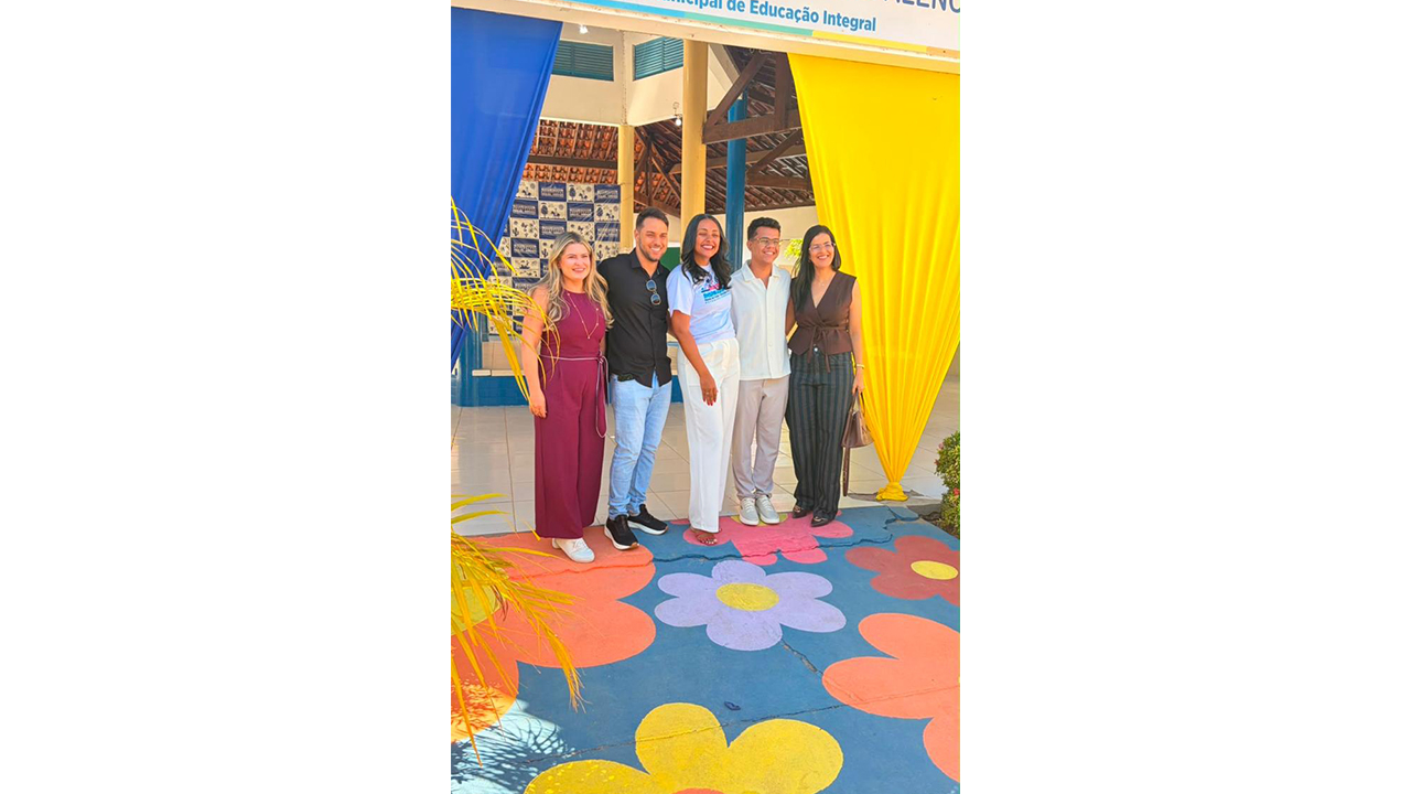Grupo multirracial de cinco pessoas posa em frente a uma escola, em pé, lado a lado, vestindo roupas casuais. À sua frente, há um piso azul pintado com grandes flores coloridas. Atrás delas, tecidos decorativos em azul e amarelo enfeitam o vão de entrada, logo abaixo de uma placa que exibe "PMEI - Política Municipal de Educação Integral". Lê-se parcialmente o nome da escola Miguel Arraes.