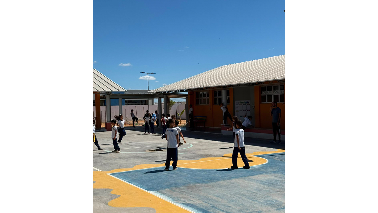 Em um pátio escolar aberto, sob um céu azul, cerca de quinze crianças correm e brincam espalhadas pelo espaço, vestindo blusas brancas e calças azuis, enquanto um adulto, em pé, as observa no lado direito. O piso da quadra é de cimento, com contornos pintados em azul e amarelo. O cenário ao fundo revela construções com paredes laranjas, janelas gradeadas e telhados brancos.