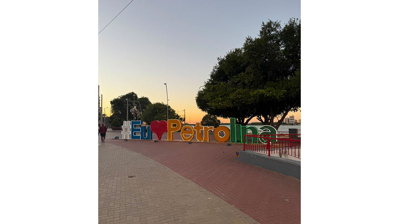 Letreiro turístico em três dimensões, apoiado no chão, onde se lê "#Eu", um símbolo de coração, "Petrolina", em amarelo, verde, branco e azul, sobre um calçadão de blocos vermelhos e cinzentos, durante o pôr do sol. Ao fundo, há árvores grandes, uma mureta com grades vermelhas à direita e a extensão de um rio.