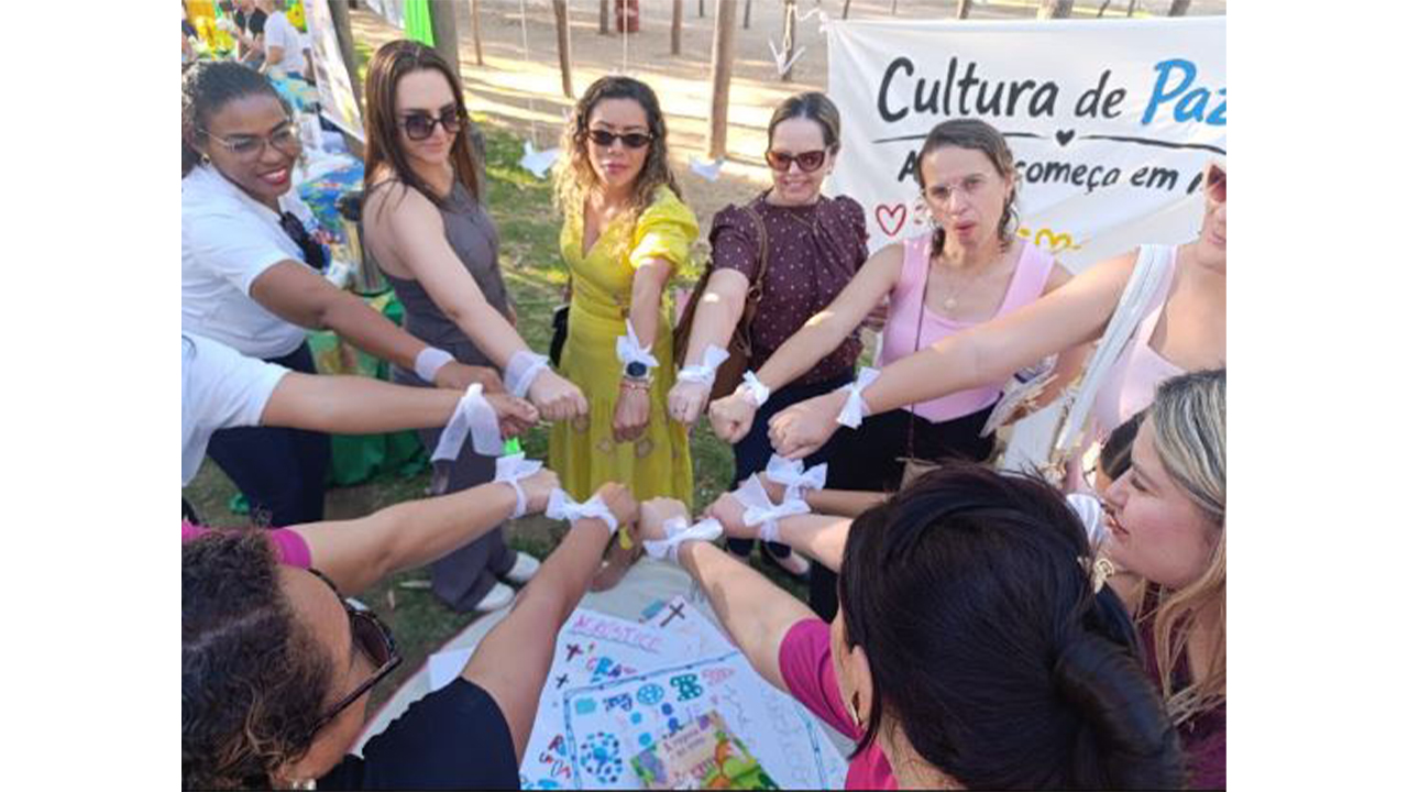 Grupo multirracial de 11 mulheres e uma criança reunidas em círculo, estendendo os braços e unindo as mãos fechadas em punho ao centro. No braço de cada uma delas, há uma fita branca amarrada na altura do pulso. Ao fundo, há um cartaz com o texto “Cultura de Paz”, árvores e materiais de exposição. Abaixo delas, no centro do círculo, é possível ver cartazes e cartolinas brancas desenhadas no chão.