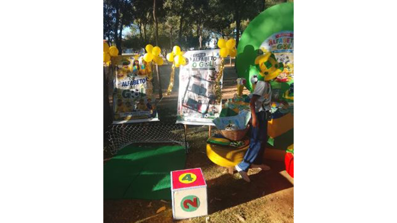 Espaço educativo ao ar livre, decorado em verde e amarelo, com painéis, balões e materiais lúdicos. Uma pessoa, aparentemente um estudante, vestindo calça jeans e um cartola de pelúcia verde e amarela, está próxima à estrutura central, onde aparecem referências a um projeto de alfabetização, com o títutlo "Do alfabeto ao gol". Ao fundo, há árvores e outros expositores. O chão é de terra batida e há um dado grande de papel com os números 4 e 2 à mostra, ao lado de um mini gol e um pedaço verde de feltro simulando um campo de futebol.