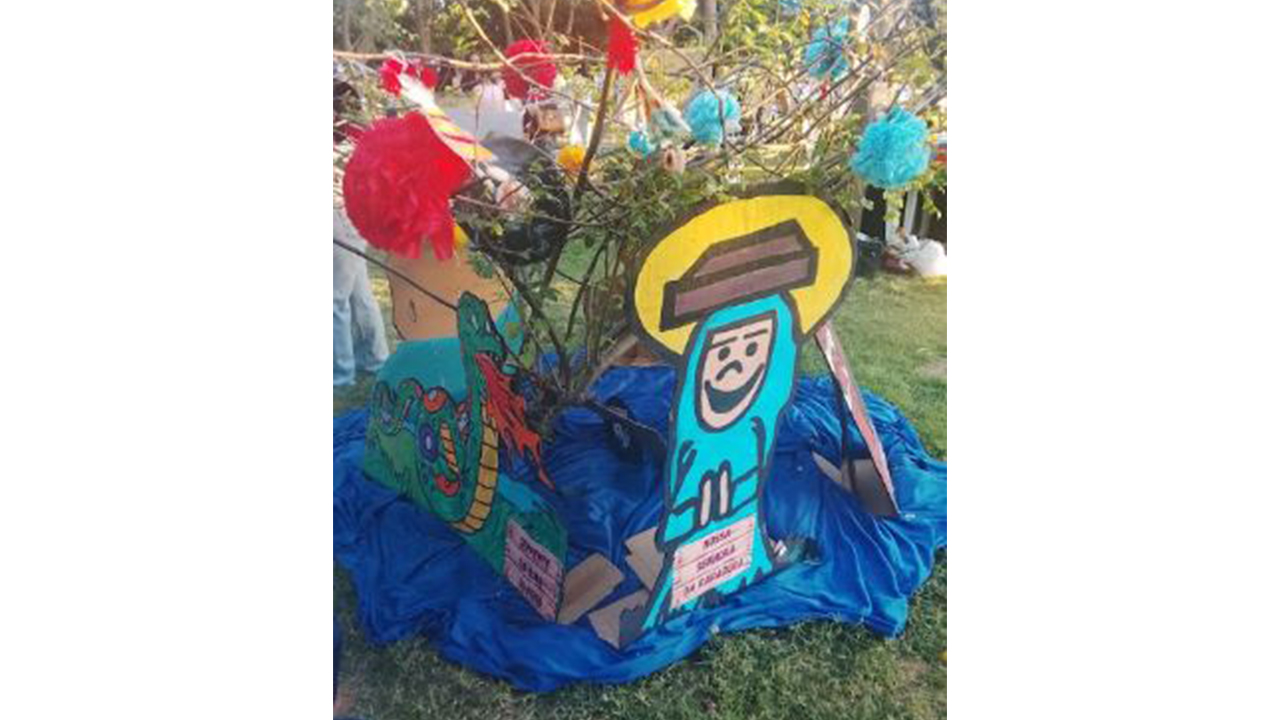 Instalação educativa montada sobre gramado, com tecido azul, galhos e enfeites coloridos suspensos. Em primeiro plano, há figuras ilustradas de personagens e elementos decorativos. Ao fundo, aparecem árvores e outras estruturas da exposição.