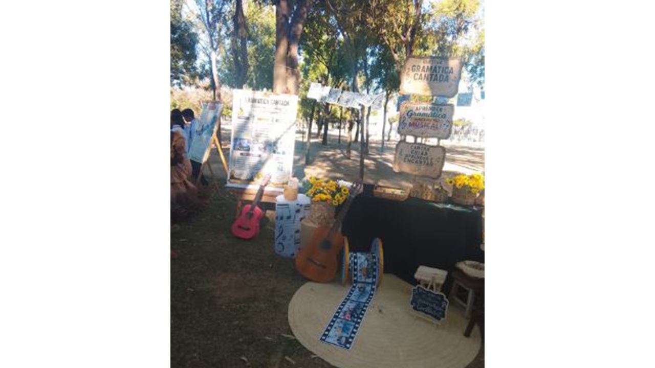 Exposição ao ar livre relacionada à música e à linguagem, organizada sob árvores. Há uma mesa com toalha preta, dois violões - um marrom e um rosa -, um carretel de filme sobre um tapete redondo de palha, cartazes, flores e materiais pedagógicos. Uma placa traz o texto “Gramática Cantada”. Ao fundo, há painéis e fotografias suspensas.