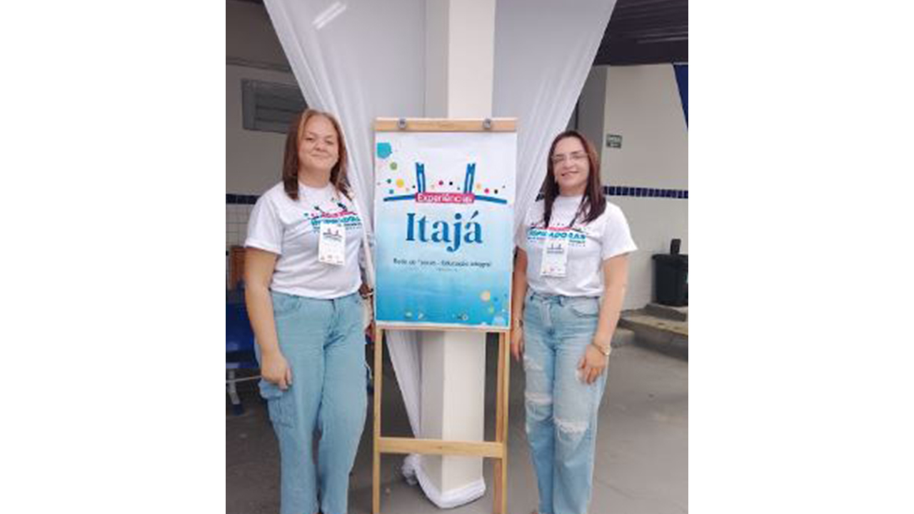 Duas mulheres, vestindo camisetas brancas e calças jeans claras, posam ao lado de um cavalete de madeira clara. O painel apresenta o nome “Itajá” escrito em azul, em um fundo degradê do branco ao azul. Ao fundo, aparecem paredes claras, um tecido branco de decoração preso a um pilar também branco, cadeiras e elementos da estrutura escolar.