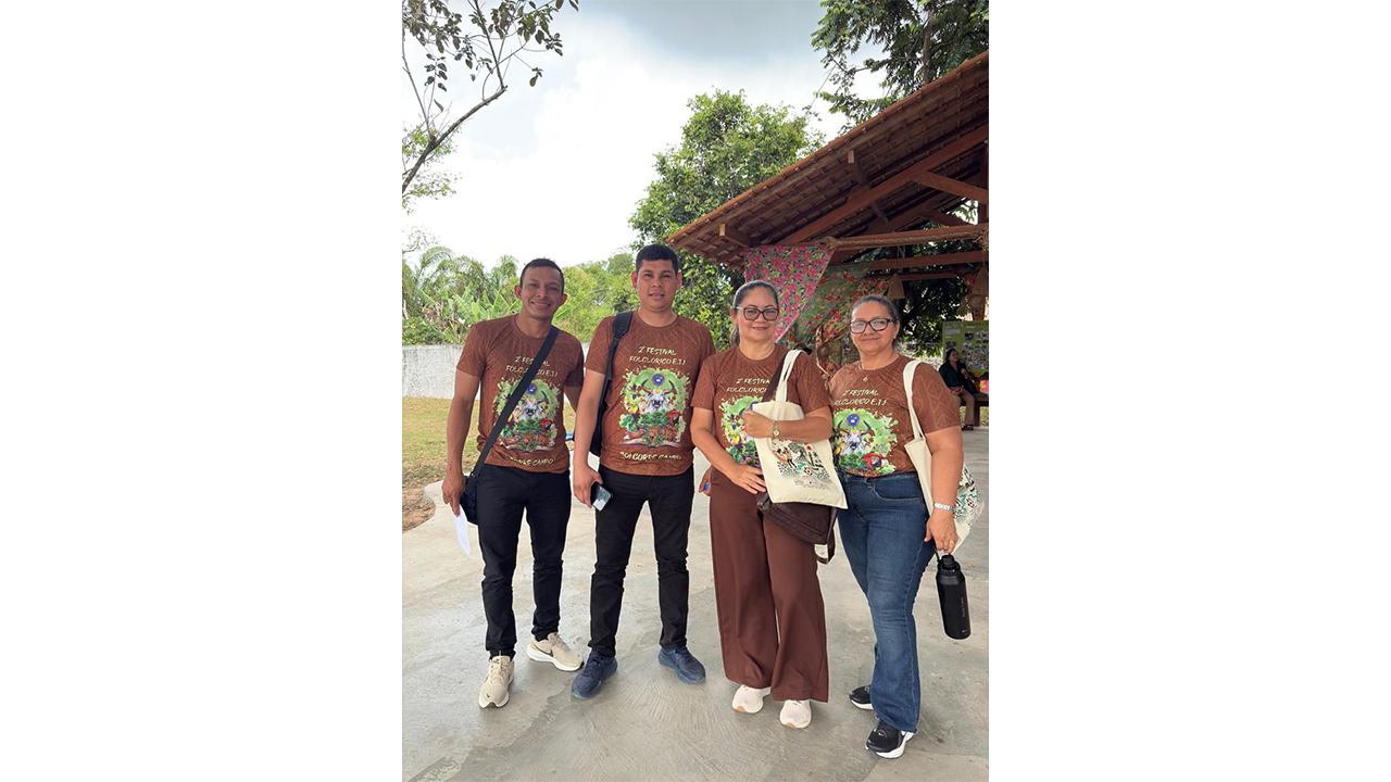 Grupo multirracial de quatro adultos posa lado a lado, vestindo camisetas marrons com uma estampa verde e o título "2º Festival Folclórico E.T.J.". O grupo está em uma área externa arborizada, próxima a uma estrutura coberta com telhado de telhas inclinado. Ao fundo, há árvores, vegetação e tecidos florais decorativos sob o telhado.
