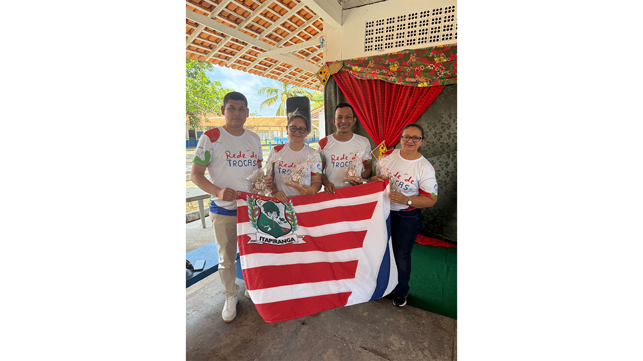 Grupo multirracial de quatro adultos posa para a foto em um espaço coberto de uma escola. Elas(es) vestem camisetas brancas com o texto “Rede de Trocas”, seguram lembranças embaladas em plástico transparente e uma bandeira com listras horizontais vermelhas e brancas e um brasão verde com a identificação “Itapiranga”. Ao fundo, há cortinas vermelha e floral, telhado de cerâmica e uma área externa sob a luz do sol.