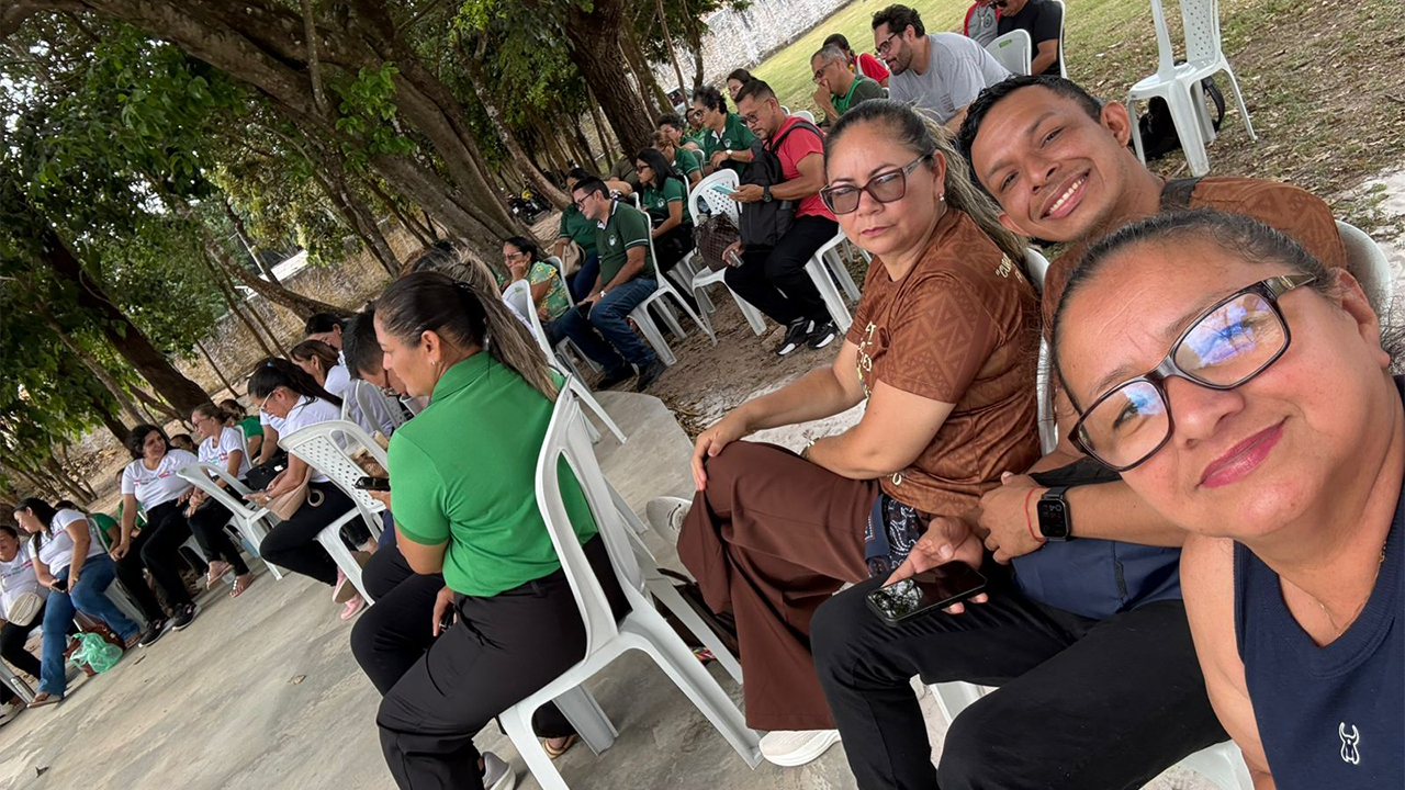 Trio multirracial de adultos posa para uma foto estilo selfie. Elas(es) estão sentadas(os) em cadeiras brancas de plástico e fazem parte de um grupo maior, de cerca de 20 pessoas, sentadas em círculo em um ambiente externo sob árvores, com piso de grama e de cimento.
