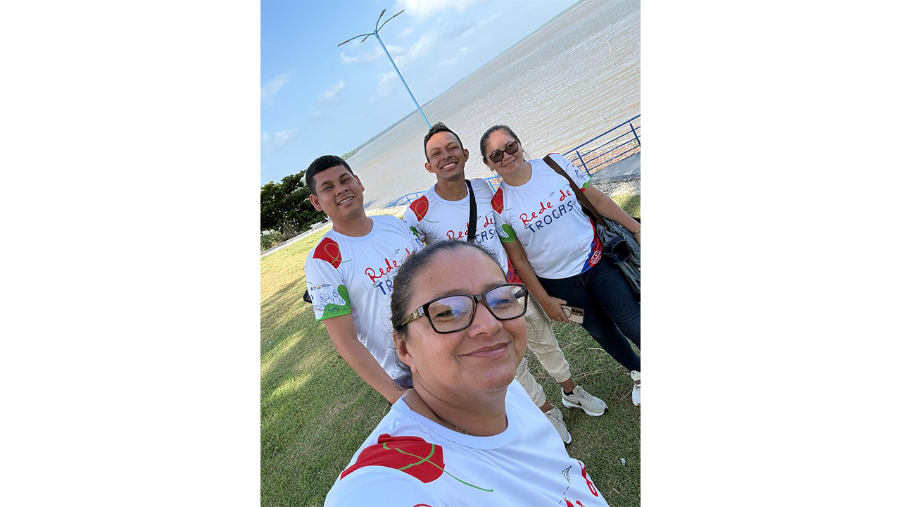 Grupo multirracial de quatro adultos tira uma selfie em pé em um gramado; a mulher à frente, usando óculos de grau, segura a câmera. O grupo usa camisetas brancas padronizadas com o texto “Rede de Trocas". Ao fundo, observam-se um caminho pavimentado, uma grade azul e uma vasta extensão de água.