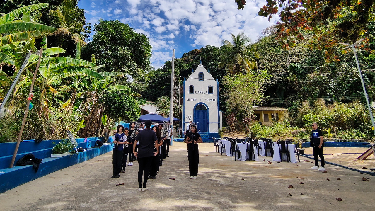 Em um pátio, um grupo multirracial de pessoas enfileiradas carrega instrumentos musicais. O destaque central é uma capela branca, com porta e contornos azuis. O cenário é cercado por árvores, bancos de concreto pintados de azul, e mesas com toalhas brancas e cadeiras pretas.