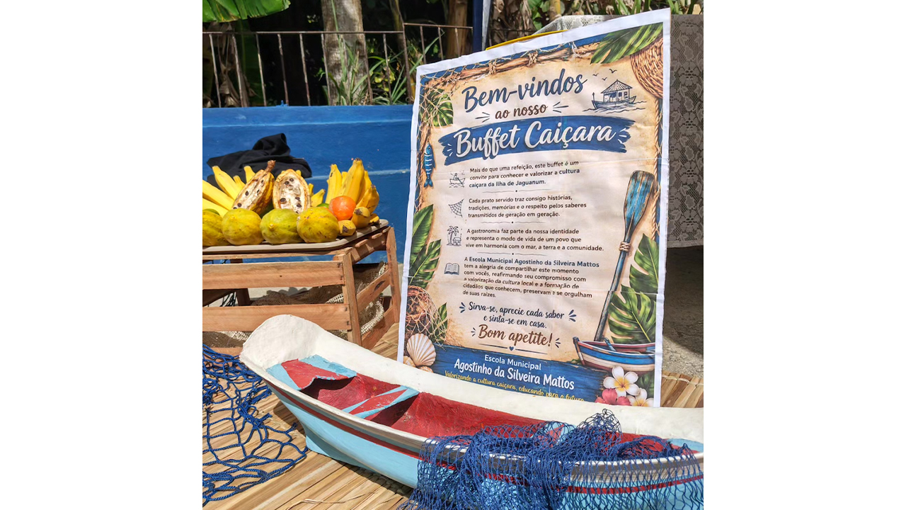 Em uma mesa, há um cartaz com a inscrição "Bem-vindos ao nosso Buffet Caiçara". Ao lado, há frutas em caixotes de madeira e uma miniatura de canoa com rede azul de pesca. O plano de fundo é composto por um muro azul e folhas verdes.