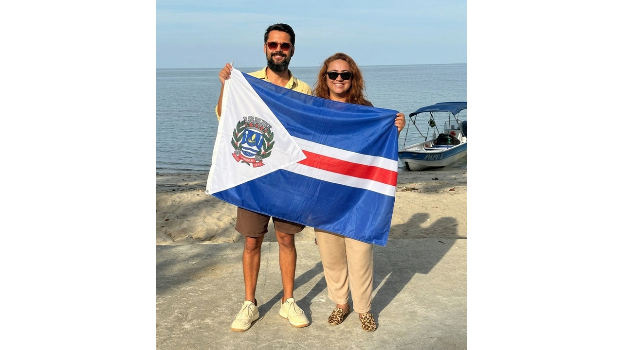 Um homem e uma mulher, ambos de óculos escuros e roupas casuais, estão em pé, lado a lado, sorrindo e segurando juntos uma grande bandeira, com faixas horizontais nas cores azul, branca e vermelha, com um brasão em um triângulo branco. Eles estão em uma faixa de areia, e, ao fundo, há o mar e um pequeno barco ancorado à direita.