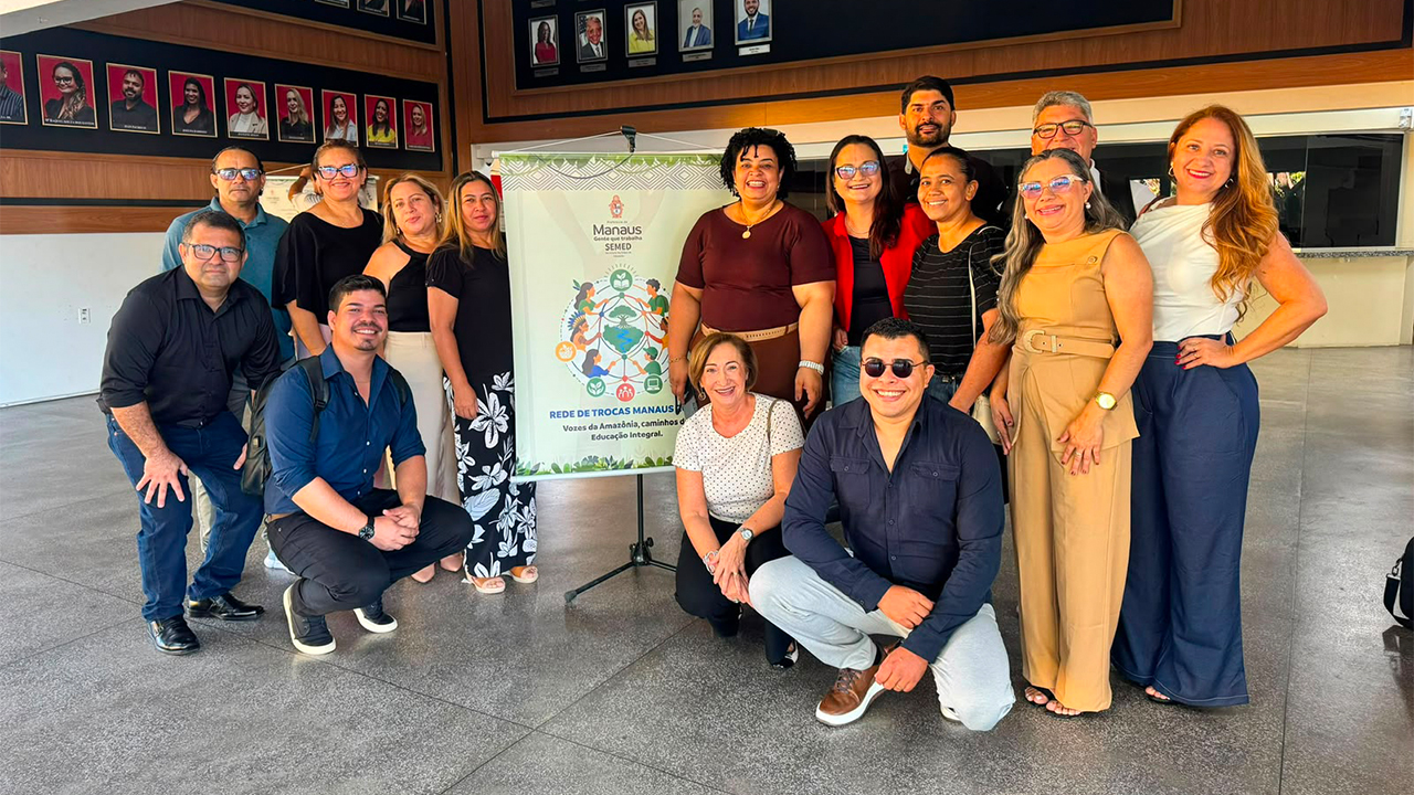 Um grupo multirracial de quinze pessoas, algumas em pé e outras agachadas, posa com roupas casuais ao lado de um tripé com banner. Ao fundo, há um piso liso e um painel de madeira escura contendo diversos retratos emoldurados.