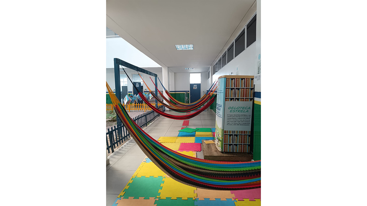 Uma fileira de redes de descanso listradas e coloridas está presa a um longo suporte de madeira. À direita, há um armário adesivado, simulando livros. O chão claro é parcialmente coberto por placas de EVA coloridas, e as paredes são brancas.