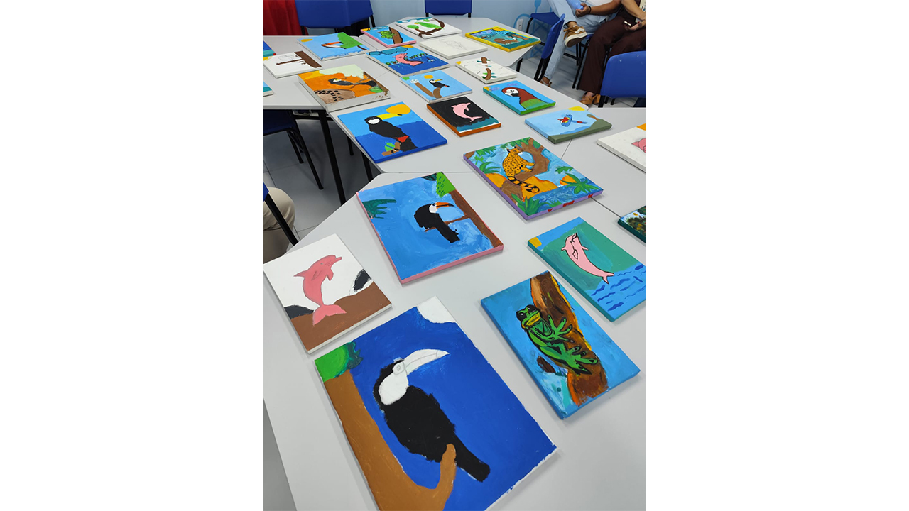 Várias telas pintadas à mão, com desenhos coloridos de animais, como tucanos e golfinhos, estão dispostas sobre uma mesa. O cenário é composto pela superfície da mesa, por cadeiras azuis dispostas ao redor e por um piso cinza liso.