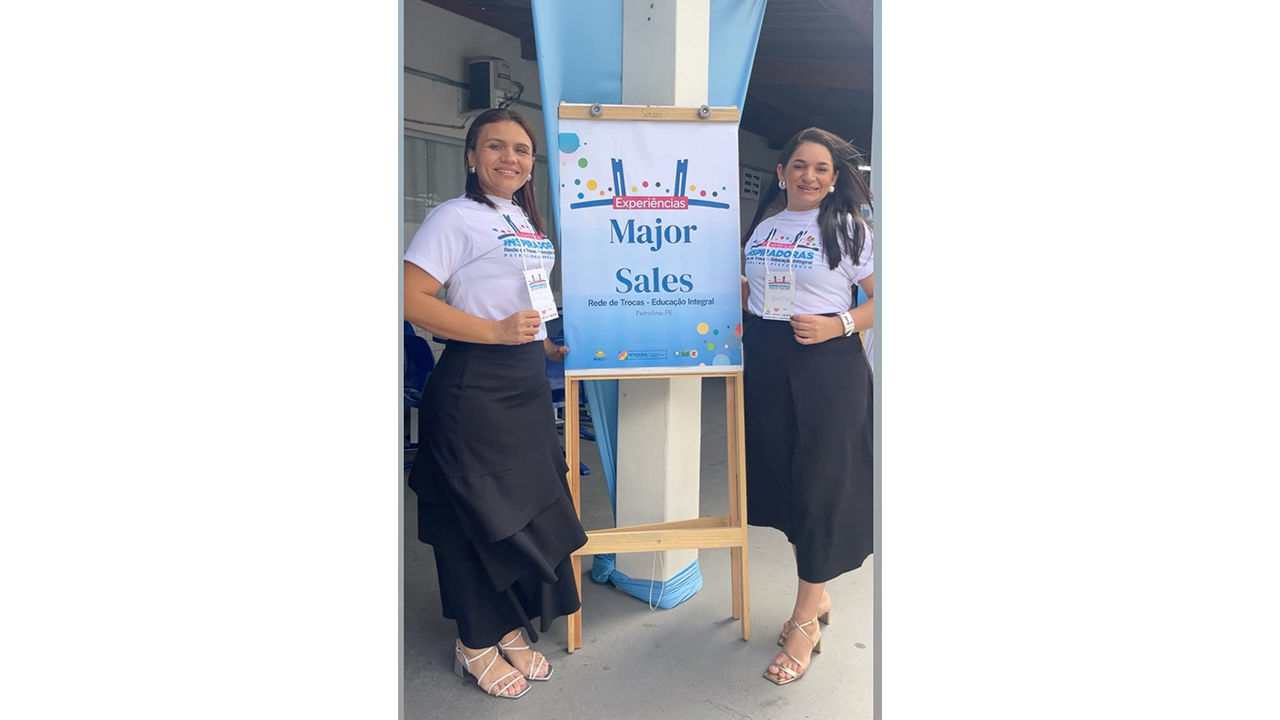 Duas mulheres estão em pé, cada uma de um lado de um cavalete centralizado, que sustenta um banner com o texto “Experiências Major Sales - Rede de Trocas - Educação Integral - Petrolina - PE”. Elas vestem camisetas brancas idênticas, com crachás no pescoço e saias pretas longas. O fundo exibe pilares brancos, adornados com tecidos azuis decorativos.