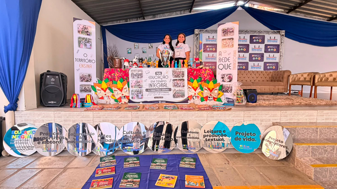 Duas mulheres, vestindo camisetas brancas idênticas, posam em pé atrás de uma mesa repleta de artesanatos, localizada em cima de um palco. Ao lado dos degraus de acesso, estão dispostos cadernos infantis sobre um tecido azul no chão, juntamente com placas redondas com temas educativos. O cenário de fundo apresenta painéis em azul e branco com a logomarca do evento, tecidos azuis pendurados em paredes brancas e um sofá bege à direita.
