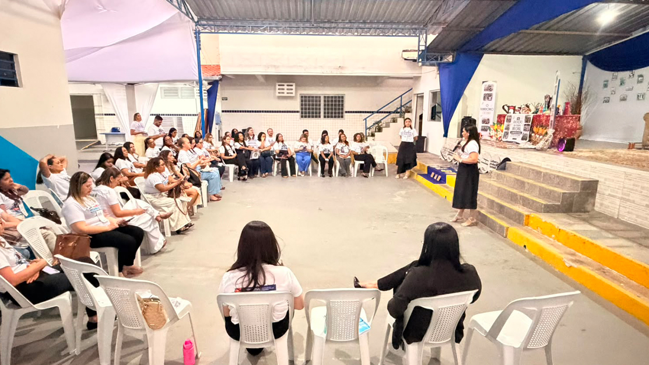Grupo multirracial de cerca de cinquenta pessoas, a maioria mulheres, está sentado em cadeiras brancas de plástico em um semicírculo. À direita, no centro do semicírculo, uma mulher de blusa branca e saia preta está em pé, segurando um microfone e falando para a plateia. O ambiente possui piso claro de cimento, telhado metálico aparente e, ao fundo, parte de um palco elevado e decorado com móveis e tecidos azuis.