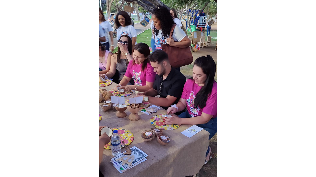 Um grupo multirracial de cerca de oito participantes está sentado ao longo de uma mesa com toalha marrom, manuseando pequenas tigelas feitas de casca de coco com o auxílio de colheres. Vestem roupas casuais, camisas brancas ou rosas. O chão é de terra, aparentemente em uma área arborizada.
