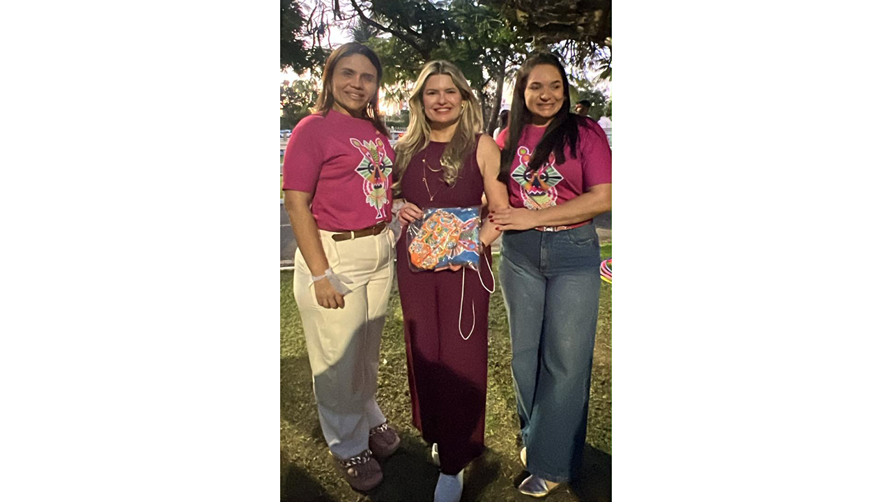 Três mulheres estão em pé, posicionadas lado a lado, sorrindo para a câmera. A mulher do centro veste um macacão vinho sem mangas, segurando, à altura da cintura, um objeto artesanal colorido, embalado em plástico transparente. As mulheres das extremidades usam camisetas rosa estampadas e calças compridas claras e escuras. O plano de fundo é composto por um gramado e pelas copas de árvores iluminadas.