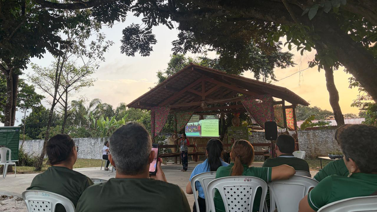 Apresentação ao ar livre sob a sombra de grandes árvores. Um grupo de aproximadamente sete adultos está sentado de costas em cadeiras plásticas brancas, voltado para uma estrutura de madeira com uma cobertura triangular com um telão projetando imagens. O espaço tem chão de cimento, um muro cinza ao fundo e árvores.