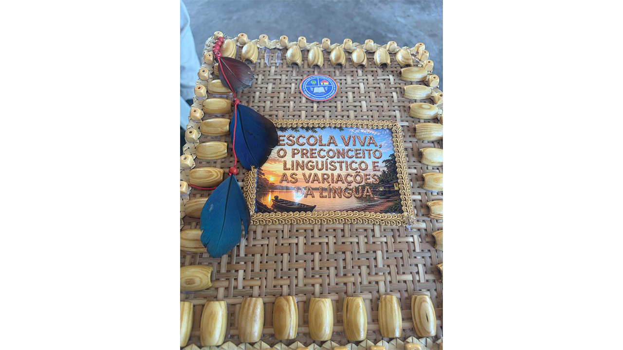 Objeto artesanal com uma capa trançada de palha, adornada nas margens com contas de madeira, penas azuis e vermelhas e um emblema na parte superior. Uma placa centralizada exibe o texto “Escola Viva, o preconceito linguístico e as variações da língua” sobre uma imagem de uma canoa em um rio ao entardecer.