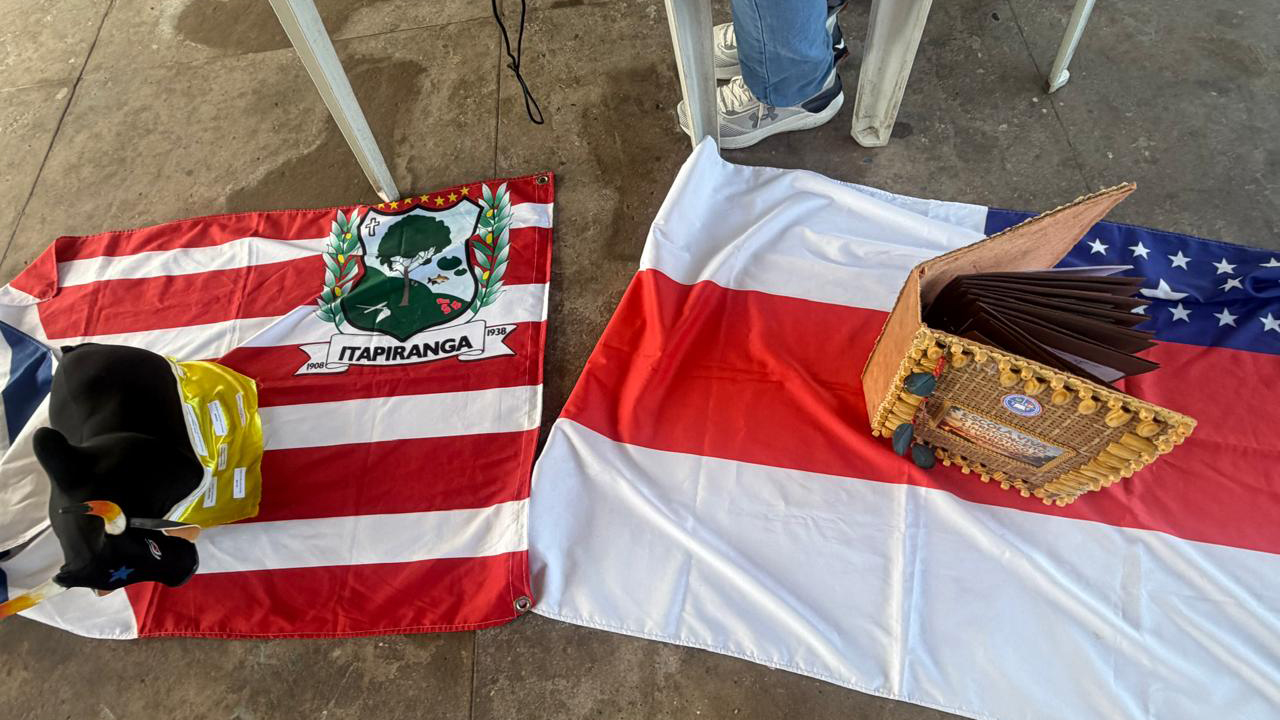 Sobre um chão de cimento, destacam-se duas bandeiras estendidas: a da esquerda, com listras vermelhas e brancas e um brasão com a inscrição “Itapiranga”, e a da direita, tricolor: branca, vermelha e azul com estrelas brancas. Sobre a bandeira da direita, repousa um livro artesanal com capa trançada, enquanto, na da esquerda, há uma miniatura de um boi preto.